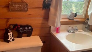 Une salle de bain rustique, aménagée dans une cabane en bois, avec des toilettes, un lavabo et une fenêtre agrémentée de rideaux.