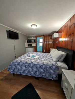 A bedroom with a bed, a television, a small kitchenette, and a door leading to another room. - Bright Seaside Cottage with Panoramic Views in North Truro, Massachusetts (North Truro)