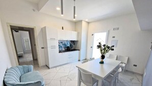 A modern kitchen with white cabinets and a marble floor, a dining area with a white table and chairs, and a light blue armchair. - At 211 Rooms Guest House - Room No. 1 (Pizzo)