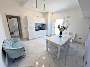 Una cocina moderna con armarios blancos y piso de mármol, un comedor con mesa y sillas blancas, y un sillón azul claro.