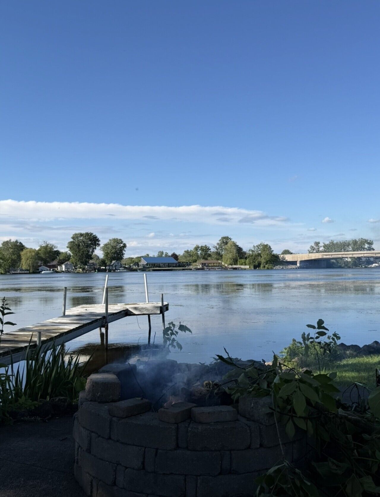 A calm lake with a dock, a stone fire pit, and a bridge in the background.