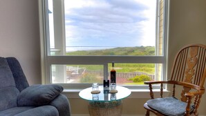 A room with a large window, a round glass table, a blue sofa, and a wooden chair. - Ocean & Jetty Views | Pool & Hot tub | #920 (Westport)