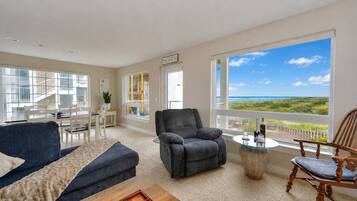 A spacious living room with a view of the beach, a gray armchair, a wooden coffee table, and a dining area with a view.