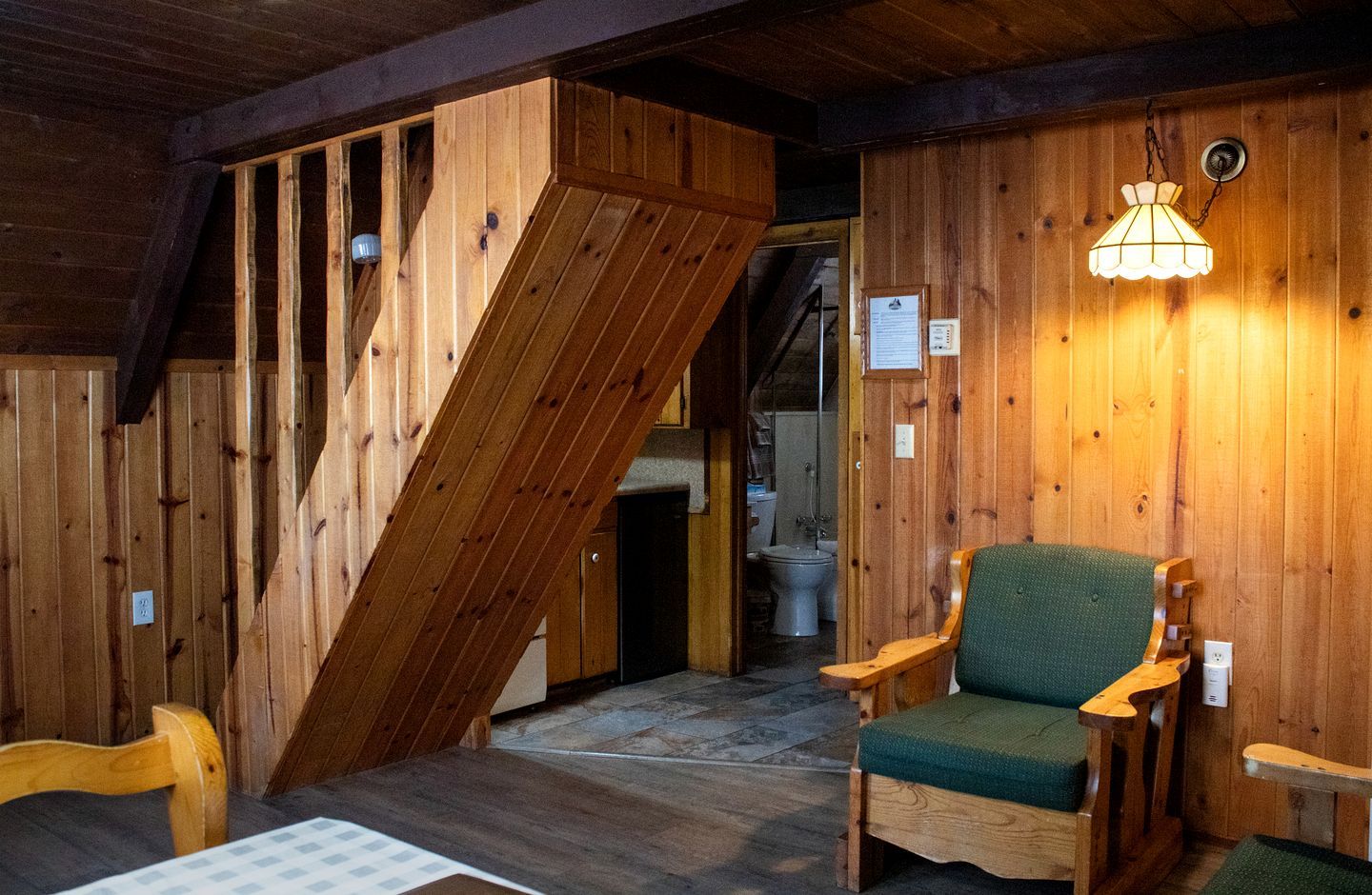 A wooden interior with a staircase, a green cushioned chair, and a hanging light fixture.