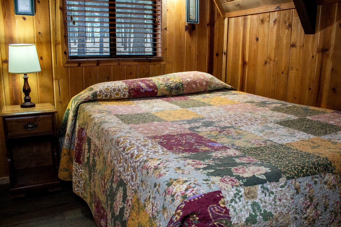 A wooden room with a bed, a nightstand, a lamp, and a window with blinds.