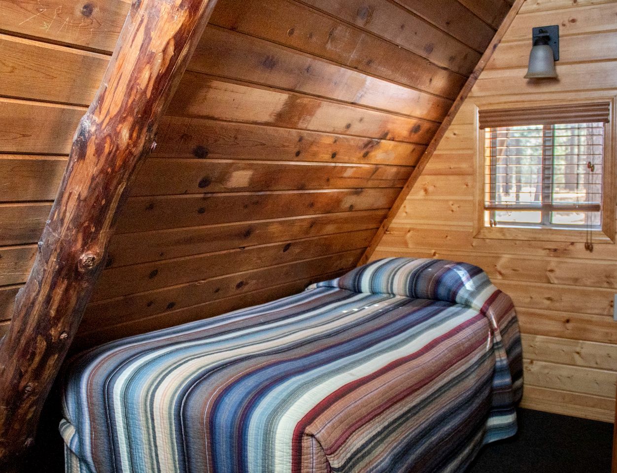 A wooden cabin room with a sloped ceiling, a bed with a striped bedspread, and a window with a view of the outdoors.