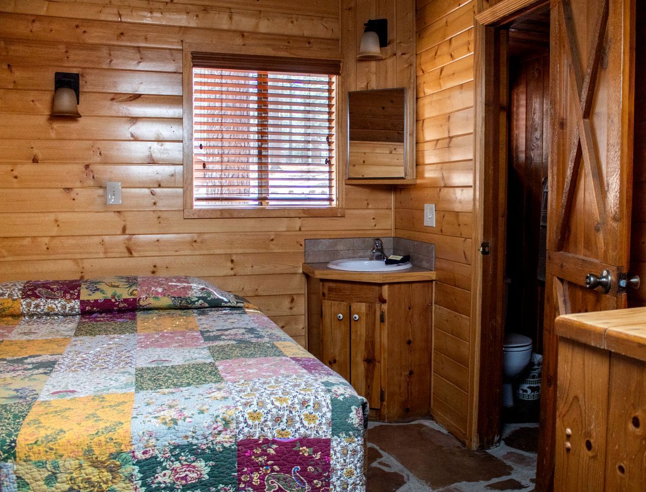 Une pièce de cabane en bois comprenant un lit, un petit lavabo et une fenêtre avec des stores.
