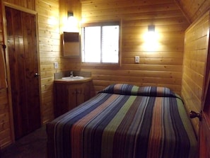 Ein Zimmer in einer Holzhütte mit einem Bett, einem kleinen Waschbecken und einem Fenster.