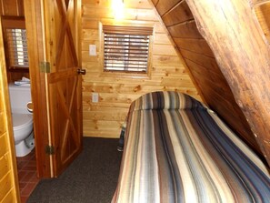 A small, cozy room with a wooden sloped ceiling, a single bed with striped bedding, a window with blinds, and a toilet visible through an open door. - Awesome Cabin Nestled Among the Towering Trees in Arizona (Flagstaff)