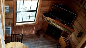 Interior de una cabaña con techo de madera, ventana, televisor, mesa de comedor con sillas y chimenea.