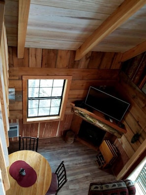 Interior de una cabaña con techo de madera, ventana, televisor, mesa de comedor con sillas y chimenea.