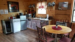 Interior de una cabaña de madera con cocina, comedor y ventana con cortinas.