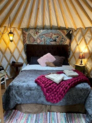 Un lit confortable dans une yourte, avec une tête de lit à motifs, un plaid rose et une petite lampe de chevet.