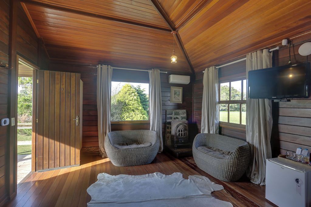 Interior de uma cabana de madeira com televisão, duas cadeiras de vime, um tapete branco e uma lareira.