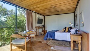 A bedroom with a large window, a bed, a bedside table, a chair, and a small desk with a TV.