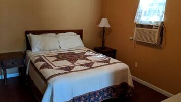 A bed with a quilt, a bedside table, a lamp, and a window with an air conditioner.