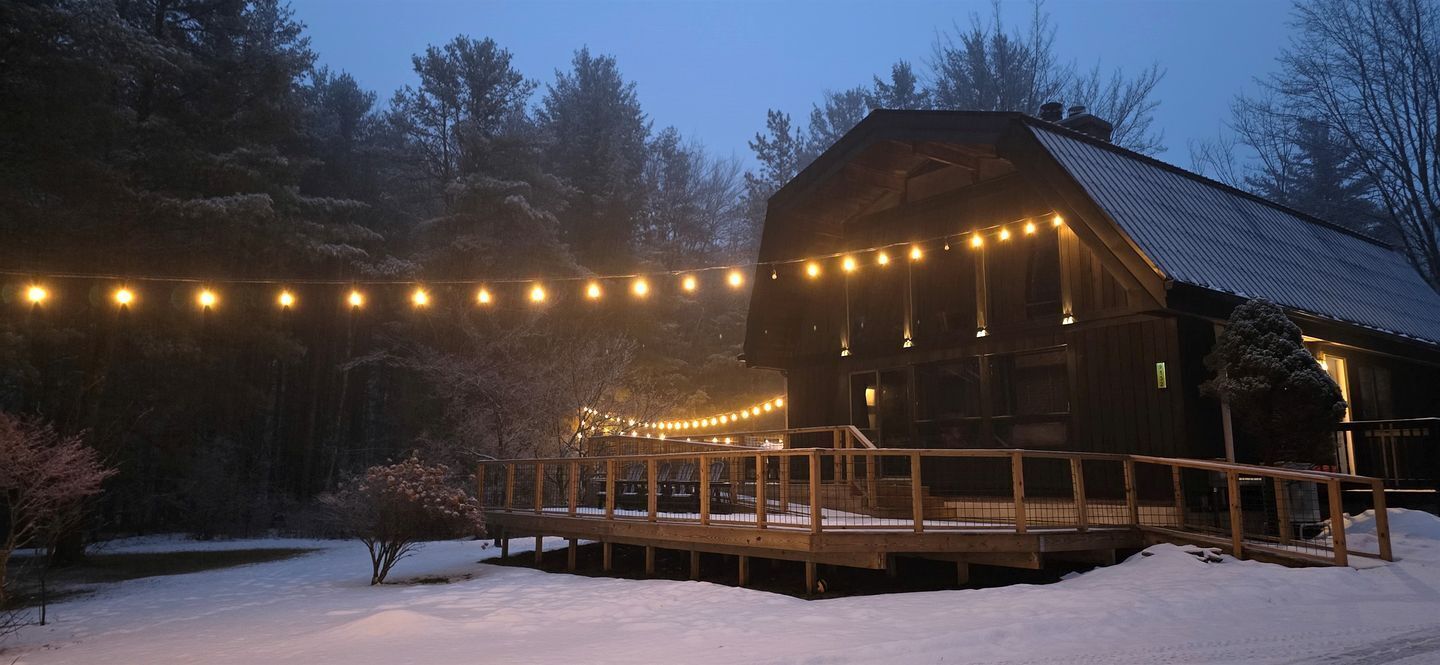 A wooden cabin with a deck, surrounded by snow and trees, illuminated by string lights.
