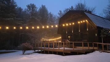 A wooden cabin with a deck, surrounded by snow and trees, illuminated by string lights.