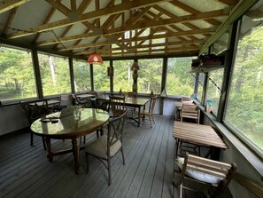 A covered outdoor dining area with wooden tables and chairs, large windows, and a view of trees.