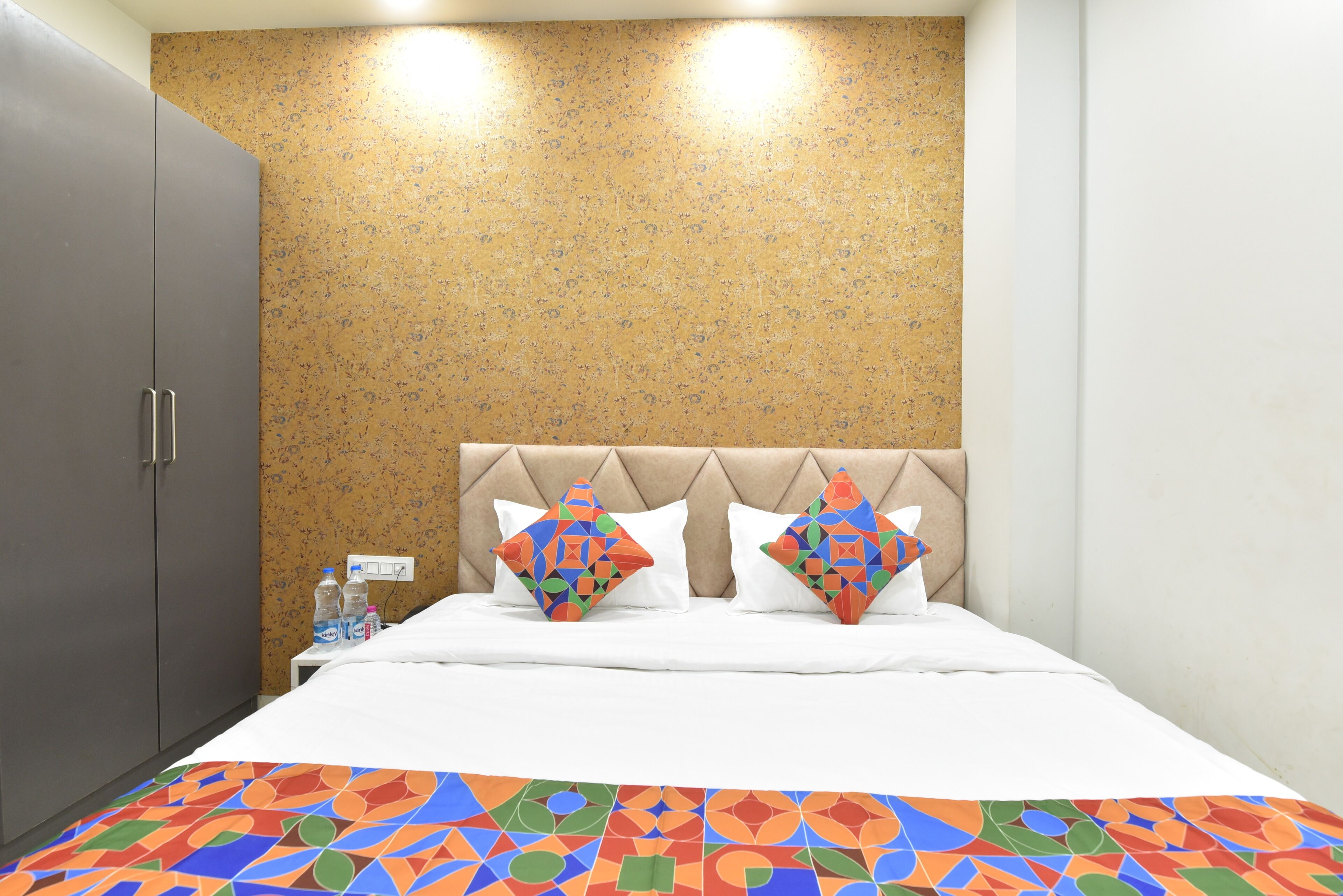 A hotel room with a bed, two pillows, a nightstand, a water bottle, and a wardrobe.