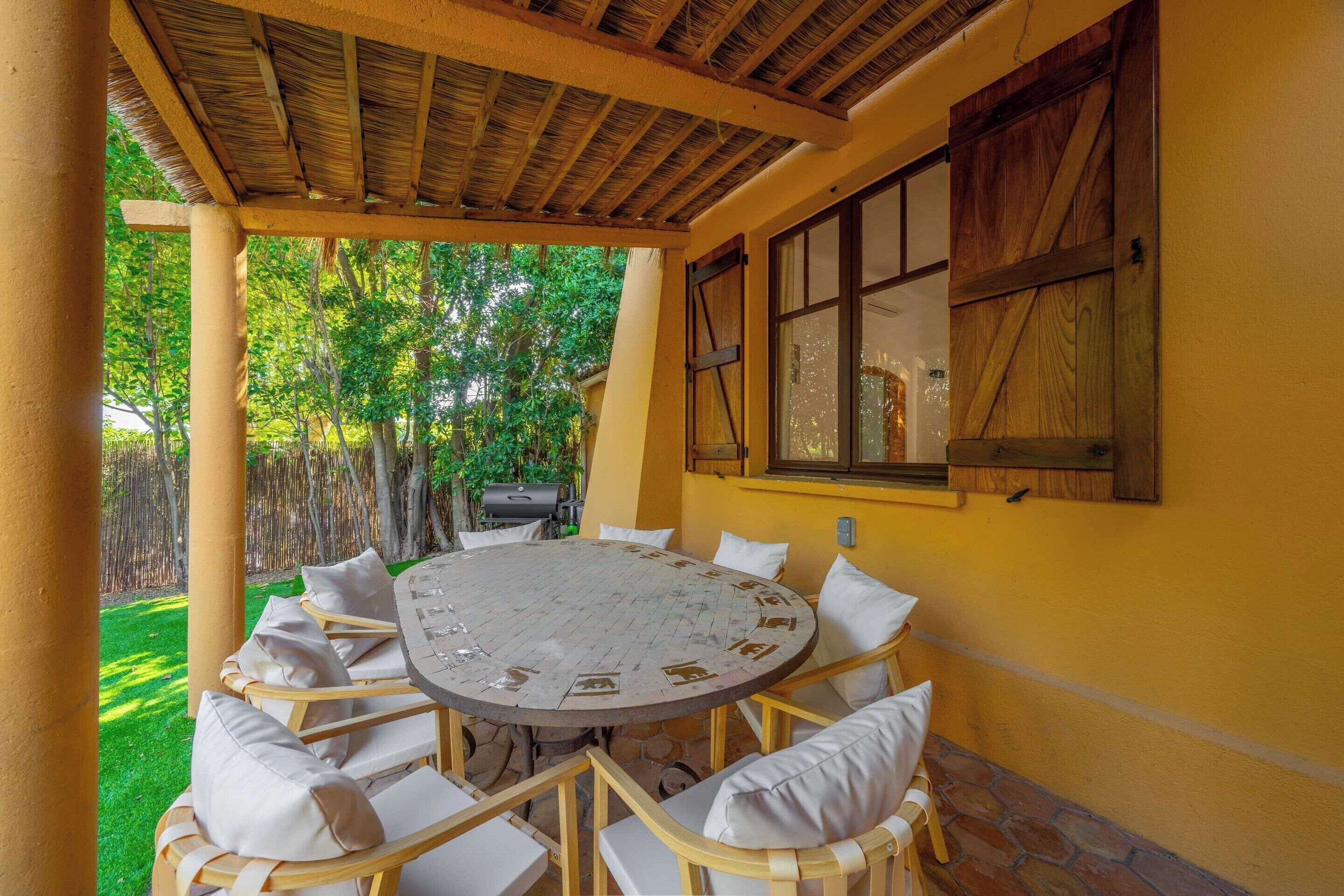 An outdoor patio with a round table, white cushioned chairs, a wooden ceiling, and a window with wooden shutters.
