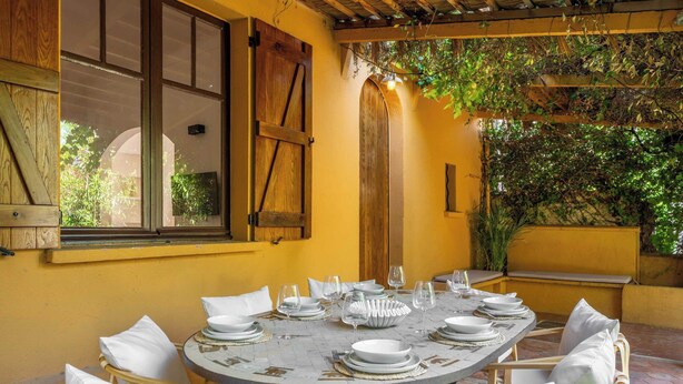 Un tavolo da pranzo rotondo apparecchiato per sei persone, con sedie bianche, un pergolato in legno e una parete gialla.