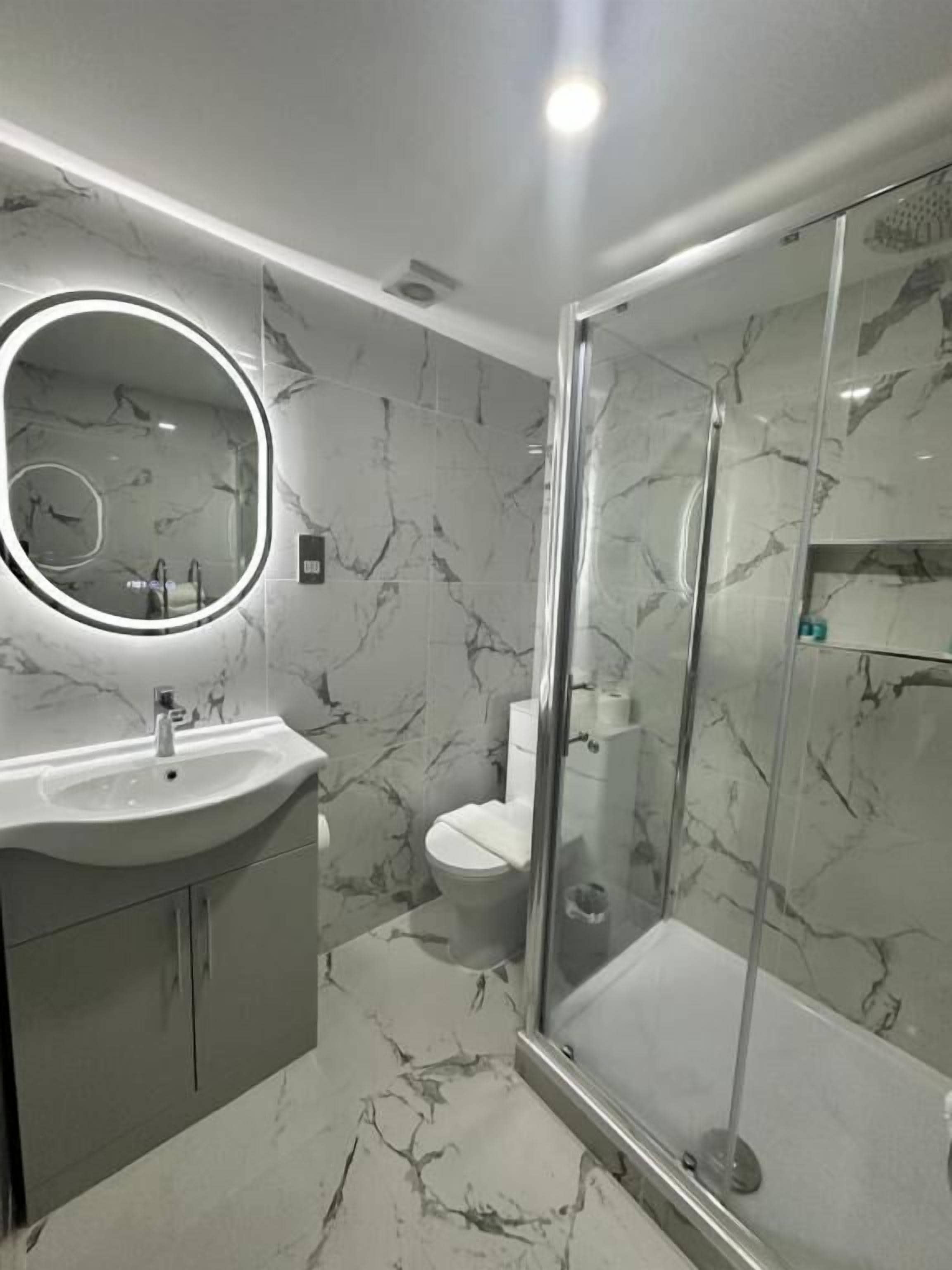 Un baño moderno con suelo de mármol, cabina de ducha de vidrio, espejo redondo con iluminación y un inodoro.