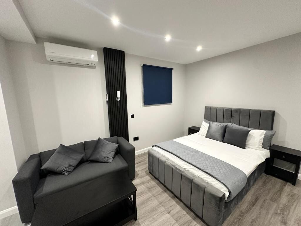 A modern bedroom with a bed, a sofa, a nightstand, and a window with blinds.