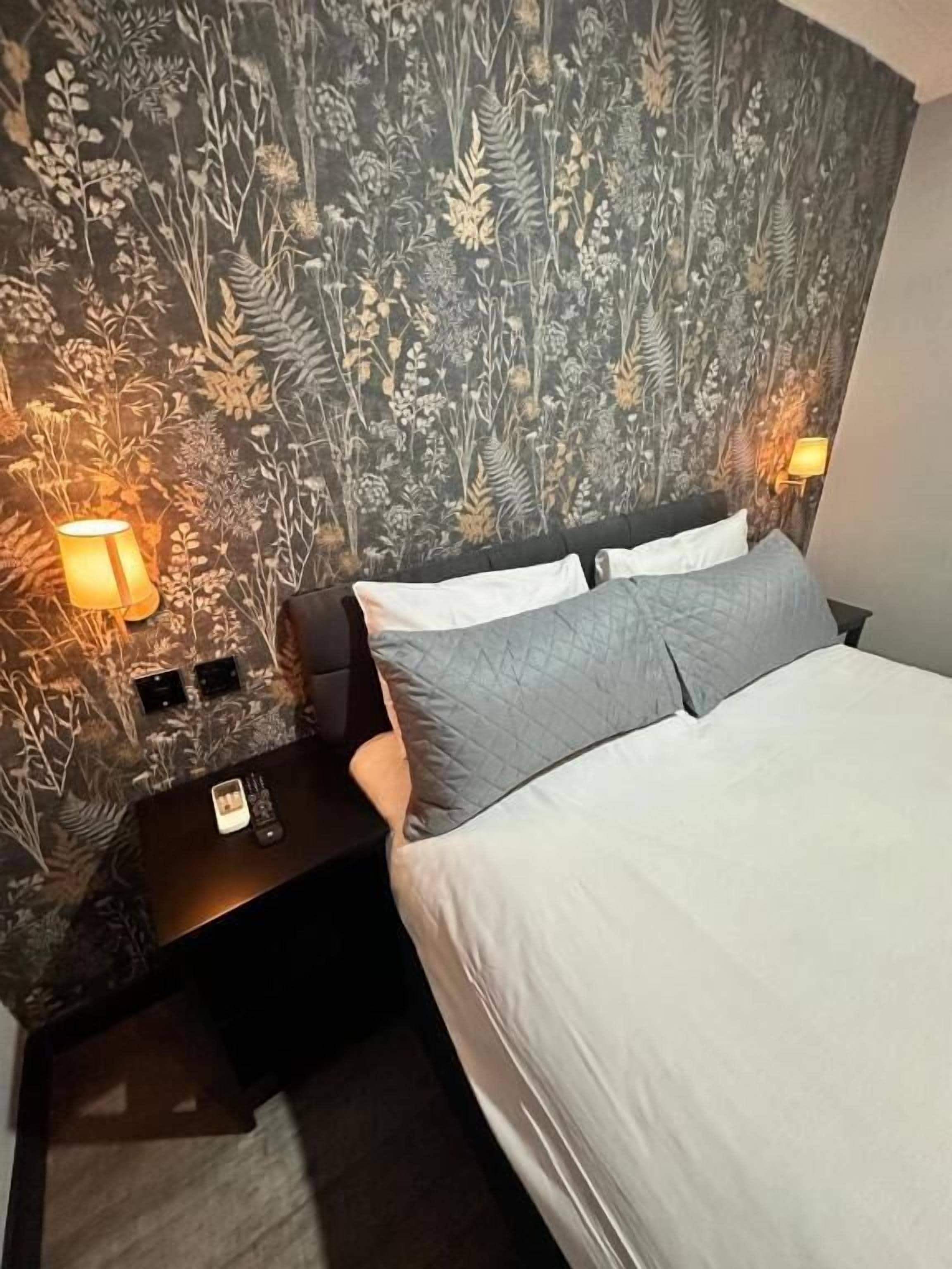 A bedroom with a patterned wallpaper, a bed with grey bedding, a nightstand with a lamp, and a power outlet.