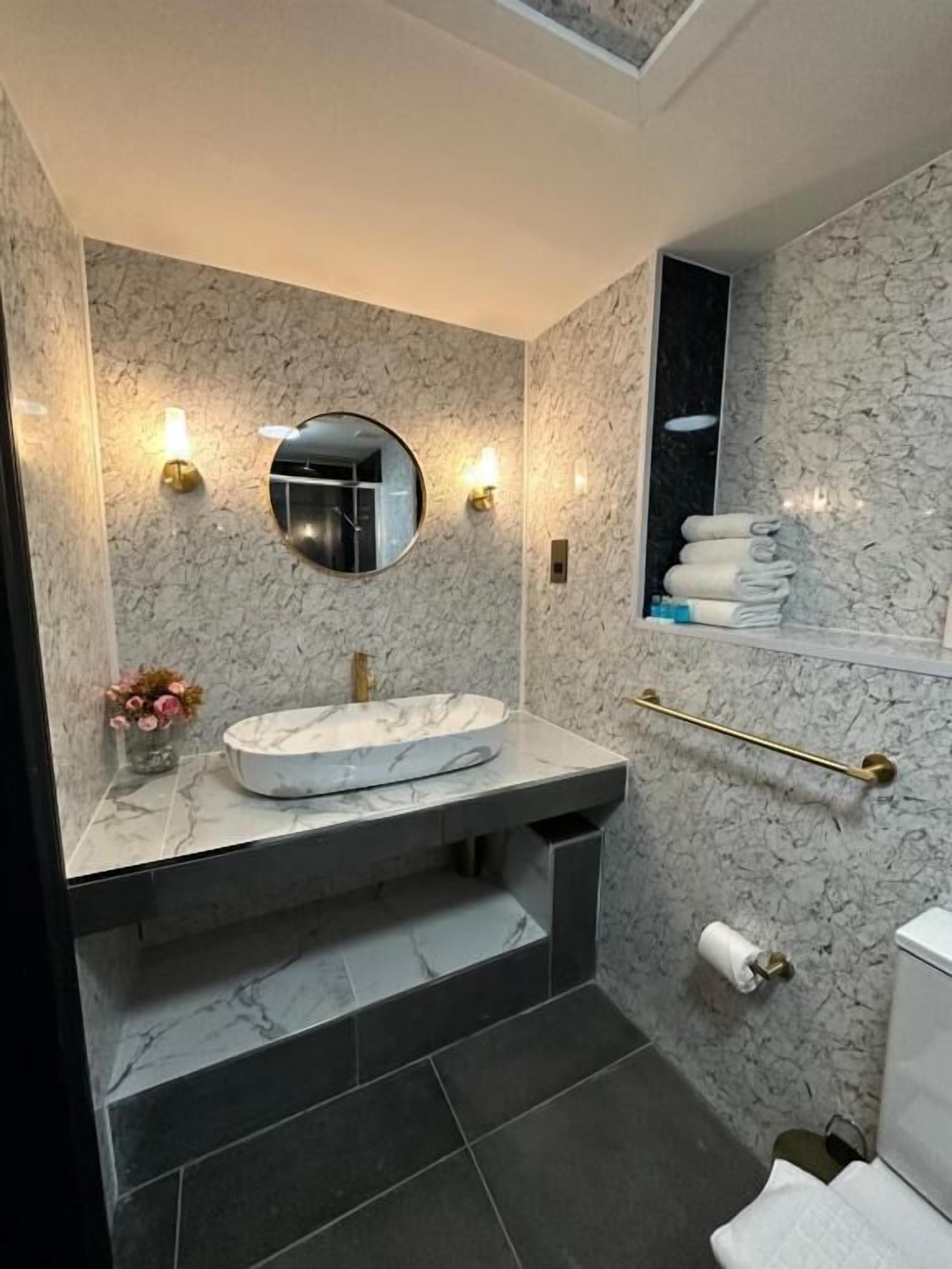 Un baño con lavamanos de mármol, un espejo redondo, apliques de pared, un perchero para toallas y un inodoro.