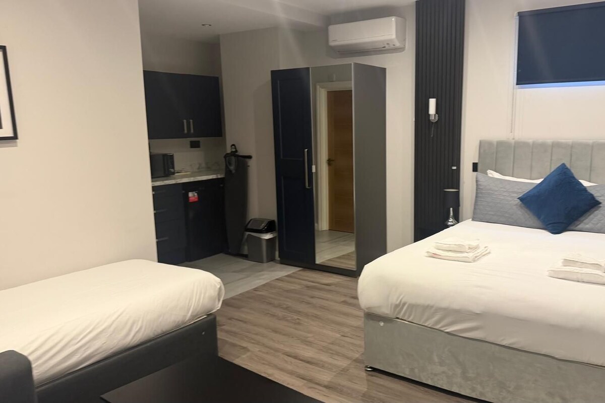 A hotel room with two beds, a small kitchenette, a mirror, and a window.