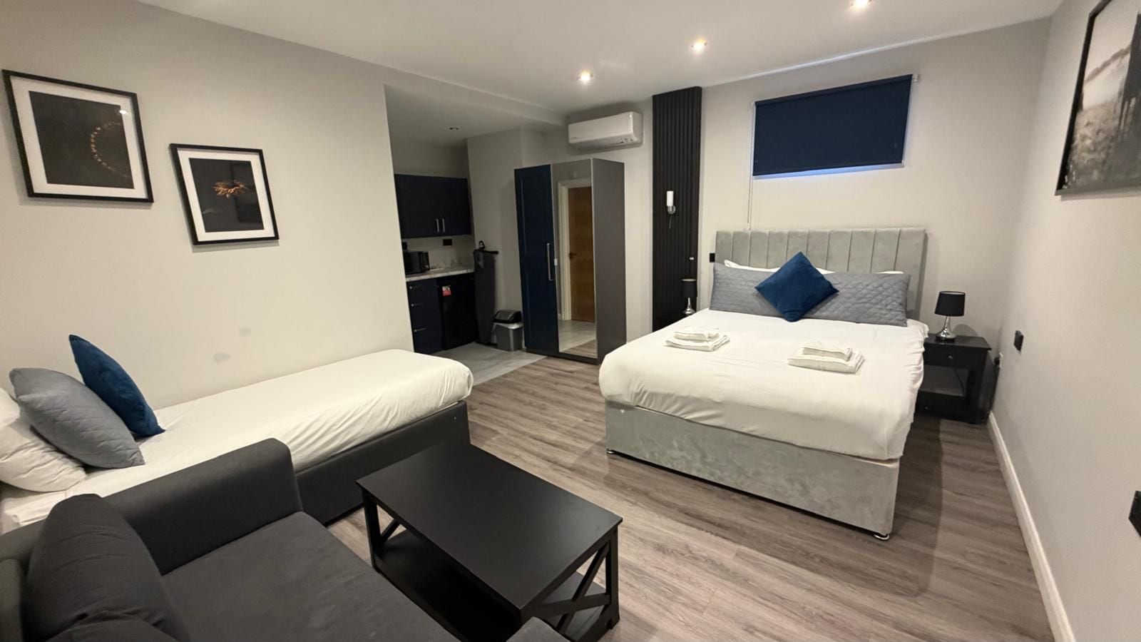 A modern hotel room with a bed, a sofa, a small table, and a desk. There is a kitchenette in the background.