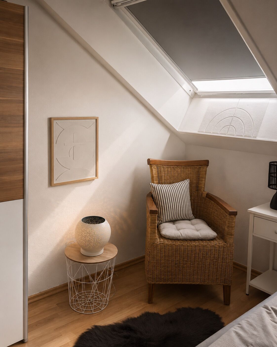 Ein gemütlicher Raum mit schrägem Dach, ein Korbstuhl mit Kissen, ein kleiner Beistelltisch mit Lampe und ein gerahmtes abstraktes Kunstwerk an der Wand.