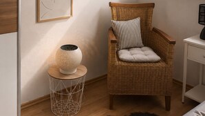 Een knus interieur met een schuine kap, een rieten stoel met kussens, een klein bijzettafeltje met een lamp en een lijst met abstracte kunst aan de wand.