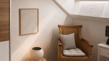 Ein gemütlicher Raum mit schrägem Dach, ein Korbstuhl mit Kissen, ein kleiner Beistelltisch mit Lampe und ein gerahmtes abstraktes Kunstwerk an der Wand.