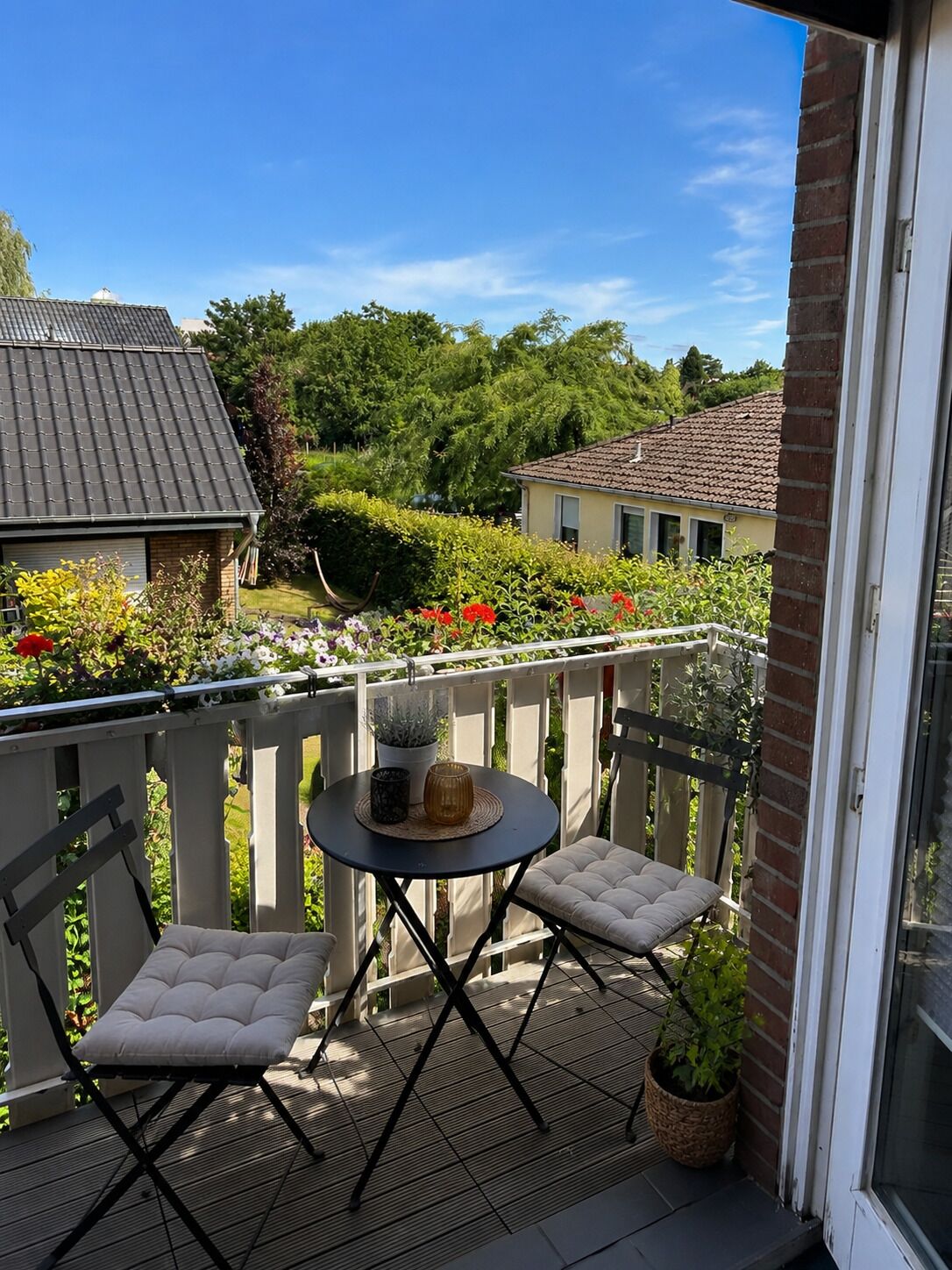 Ein Balkon mit rundem Tisch und zwei gepolsterten Stühlen, einem Blumentopf und Blick auf eine Wohngegend mit Häusern und Grünflächen.