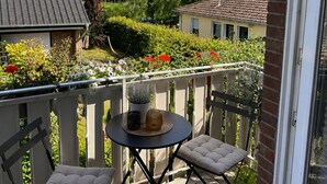 Een balkon met een ronde tafel en twee stoelen met kussens, een bloempot en uitzicht op een woonwijk met huizen en groen.