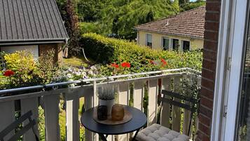 Ein Balkon mit rundem Tisch und zwei gepolsterten Stühlen, einem Blumentopf und Blick auf eine Wohngegend mit Häusern und Grünflächen.