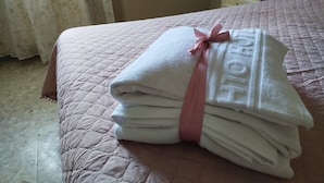 Una cama bien hecha con un edredón rosa, una pila de toallas blancas dobladas atadas con un lazo rosa, y una mesita de noche blanca.