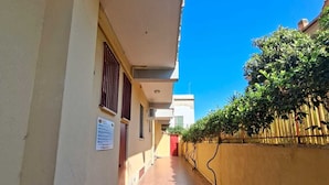 Un pasillo estrecho con suelo de baldosas, una galería techada y una pared amarilla con valla y plantas.