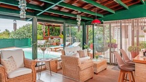 A covered outdoor seating area with wicker furniture, a glass table, and a view of the pool and greenery. - Villa Stachy & Elia with Heated Pool, by Estia (Malevizi)