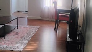 A spacious living room with hardwood flooring, a dining area with a table and chairs, a television, and a large window with sheer curtains.