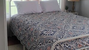 A bed with a patterned bedspread, a bedside lamp, and sheer curtains.