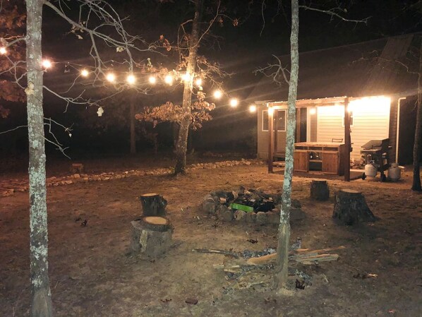 Entorno rústico al aire libre con una estructura de madera, posiblemente una cabaña o cobertizo, rodeado de árboles y un pozo de fuego.