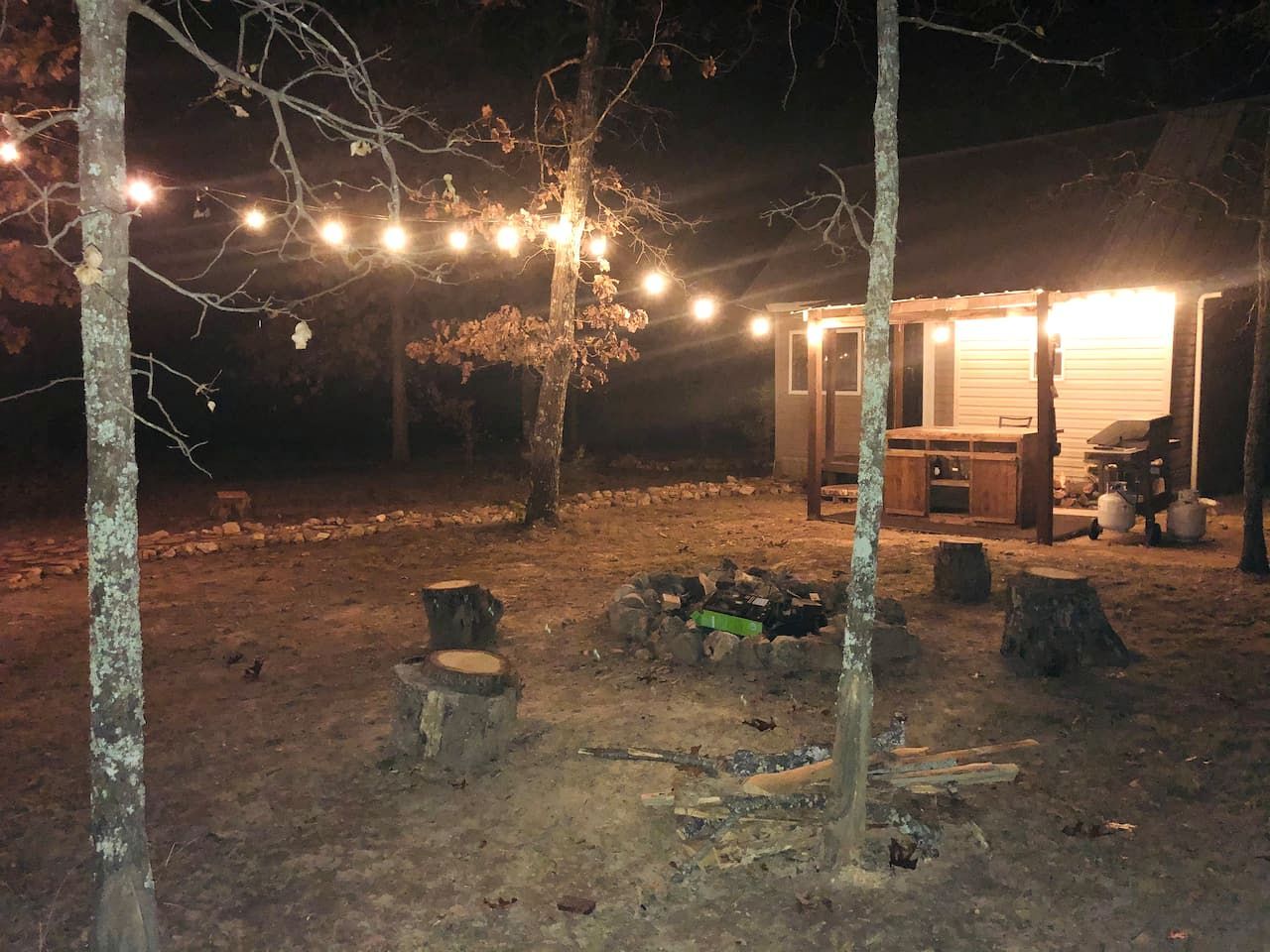 Un cadre extérieur rustique, avec une structure en bois, peut-être une cabane ou un abri, entouré d’arbres et agrémenté d’un foyer.