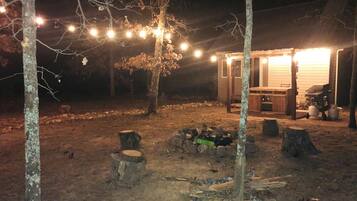 A rustic outdoor setting with a wooden structure, possibly a cabin or shed, surrounded by trees and a fire pit.