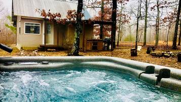 Un jacuzzi con terraza de madera, una pequeña construcción con porche y árboles al fondo.