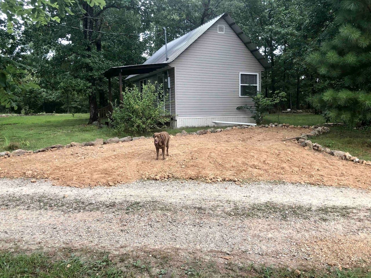 Una casa con porche cubierto, patio cercado y un perro de pie sobre el camino de grava.