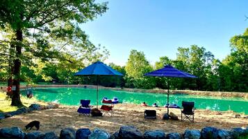Zona arenosa de playa con dos sombrillas azules, una piscina y varias sillas.