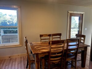 A dining area with a wooden table and chairs, a window with a view of trees, and a door with a view of a balcony. - Spacious Cabin with Fire Pit, Perfect for a Family Getaway to Custer State Park (Sturgis)